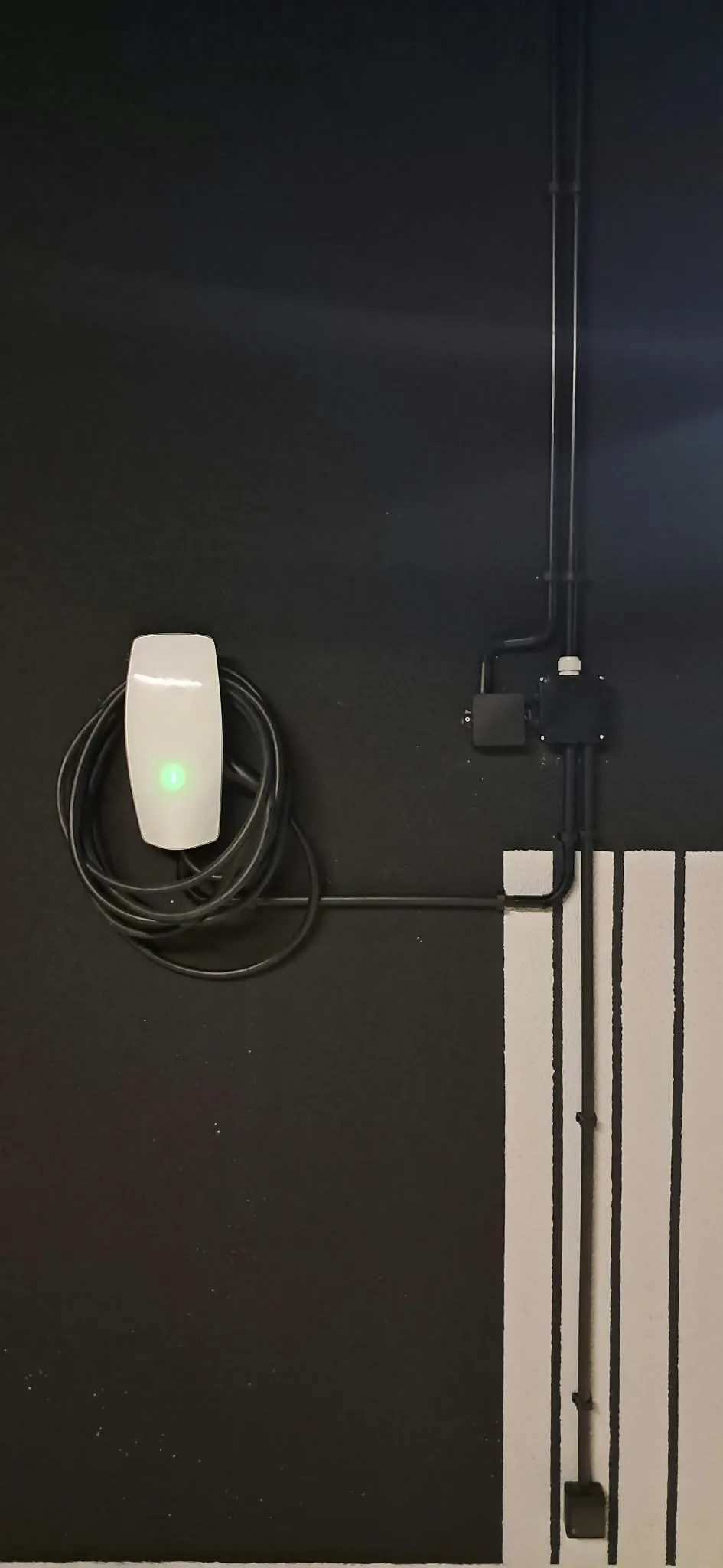 Biała ładowarka Tesla Wall Connector 11 kW z zieloną diodą i zwiniętym kablem na czarnej ścianie garażu podziemnego, rury instalacyjne natynkowe i puszki, Powiśle Warszawa